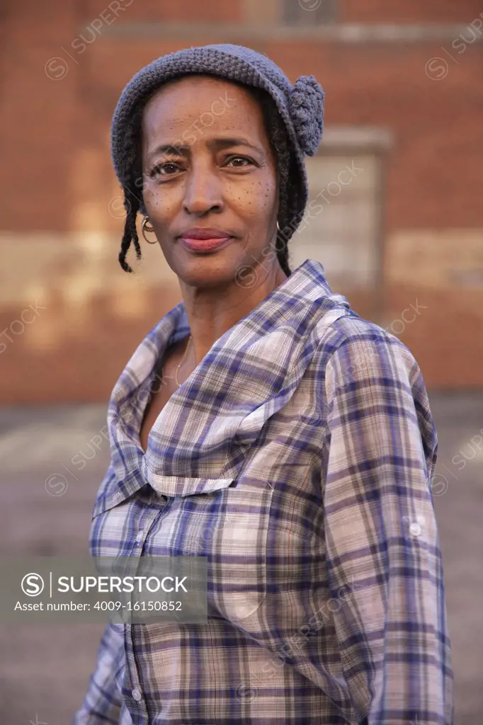 Portrait of older woman wearing knit hat standing in alley, brick wall in background  