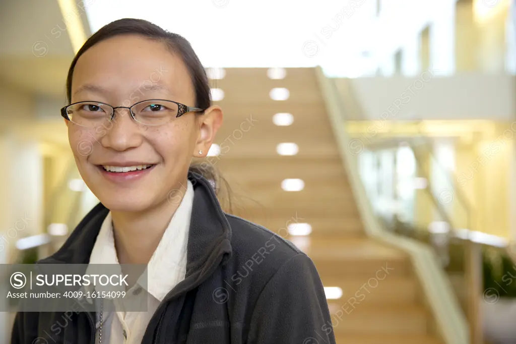 Portrait of young ethnic woman wearing glasses standing by stairwell in lobby of building, smiling looking towards camera 