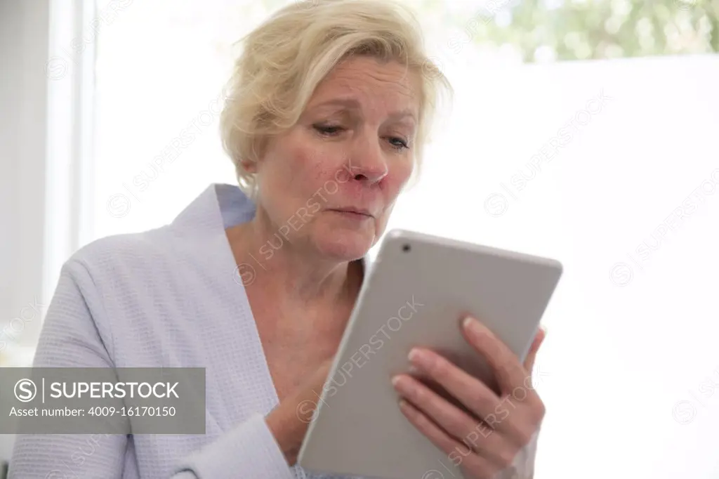 Sick senior Caucasian female sitting on side of bathtub getting ready to fill it for a bath, feeling ill using tablet to get health advice from doctor 