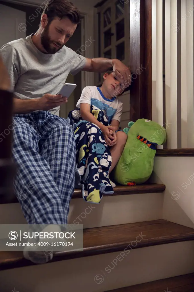 Father and son sitting at top of stairs in home getting ready for bedtime , dad feeling for fever and checking cellphone 