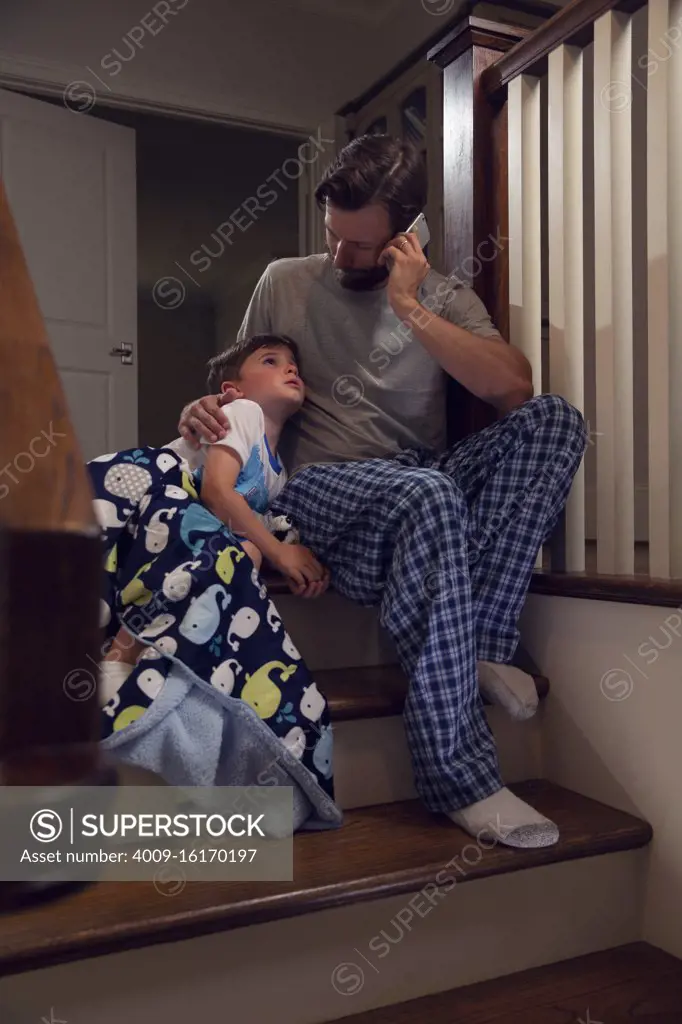 Father and son sitting at top of stairs in home getting ready for bedtime, dad calling doctor and checking on his son who is feeling ill 