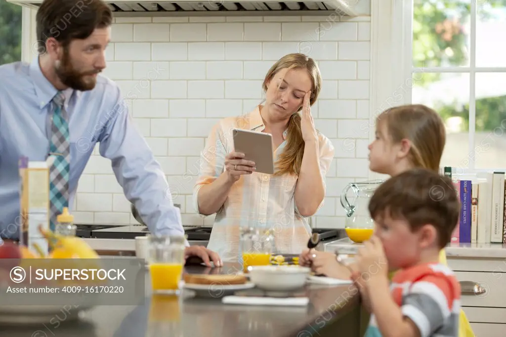 Mom with family in kitchen, struggling to get the kids breakfast and ready to start the day. Checking tablet to connect with doctor and ask about her headaches
