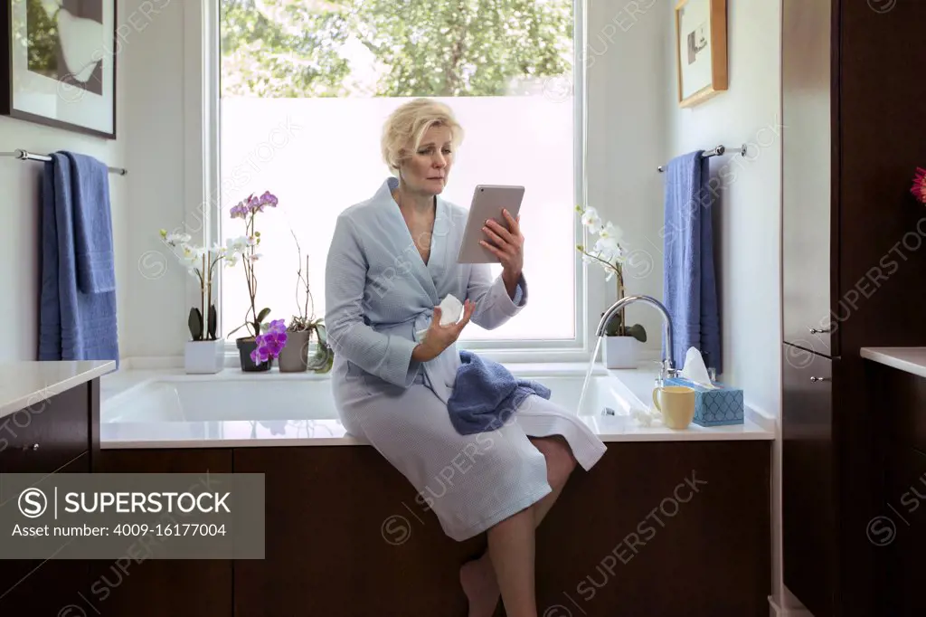 Sick senior Caucasian female sitting on side of bathtub getting ready to fill it for a bath, feeling ill using tablet to get health advice from doctor 