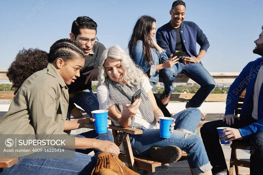 Group of young co-workers hanging out on rooftop patio laughing and having a drink, sharing images on mobile devices 
