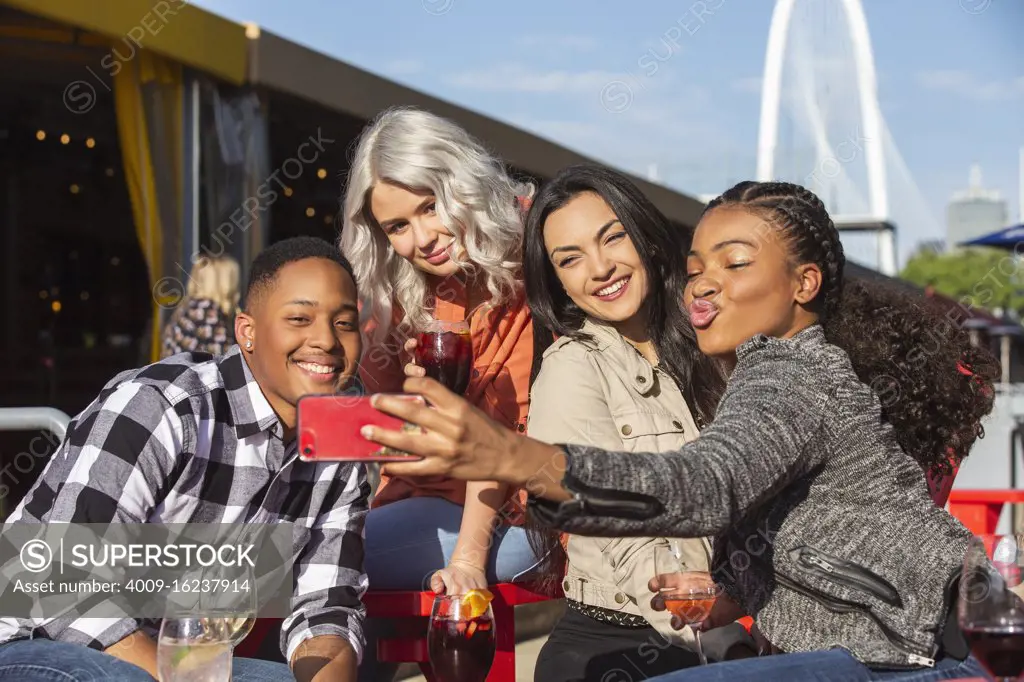 Group of young co-workers hanging out on outdoor patio having drinks for happy hour, woman taking group photo with mobile phone 
