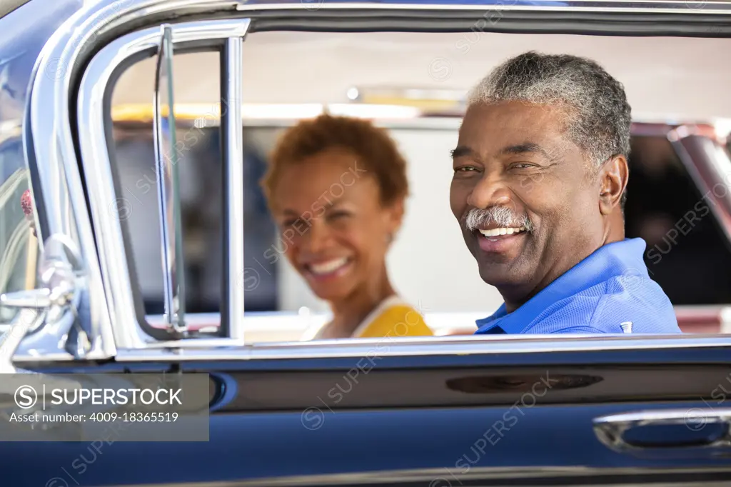 Older couple sitting in a classic car looking to camera smiling