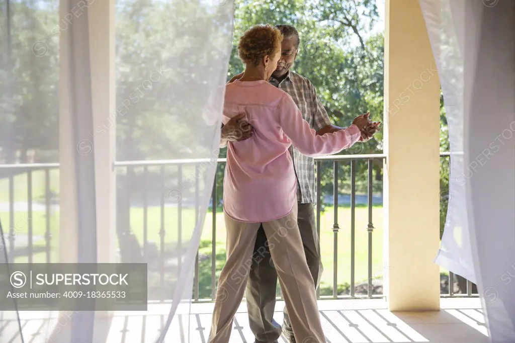 Older couple dancing on porch overlooking  green lawn , seen through sheer curtains 