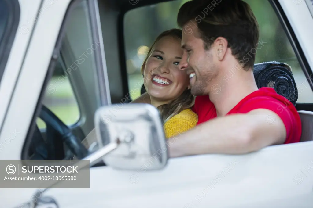 Young couple sitting in vintage pick up truck , guy with his arm around the girl while she looks over at him smiling. 