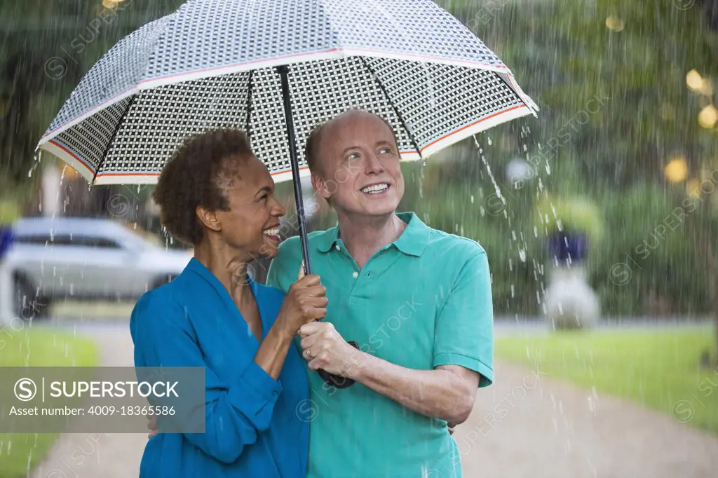 Older couple walking through park with umbrella in the rain, man looking out at the weather 