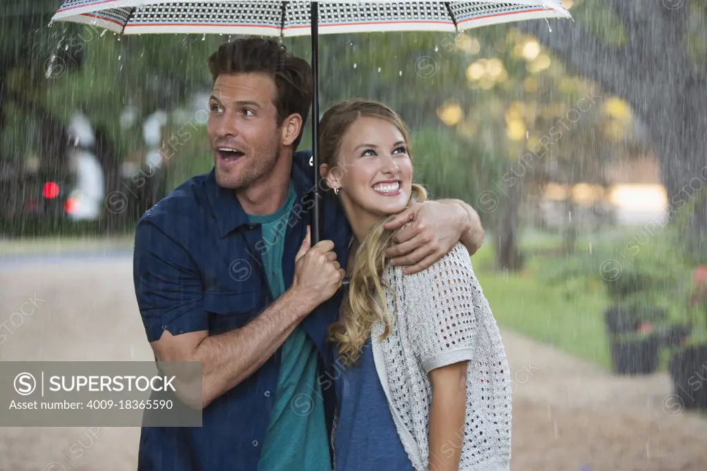Young couple walking through park with umbrella in the rain, both looking out at the falling rain 
