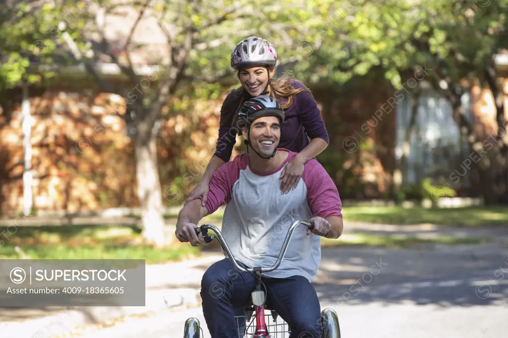 Young couple with helmets riding large tricycle through neighborhood, girl standing on back with her arms on boyfriends shoulders 