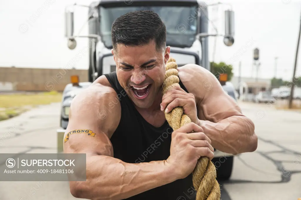 Muscular Hispanic man wearing tank top with “Strong” tattoo on his bicep, Straining to pull semi truck with large tow rope 