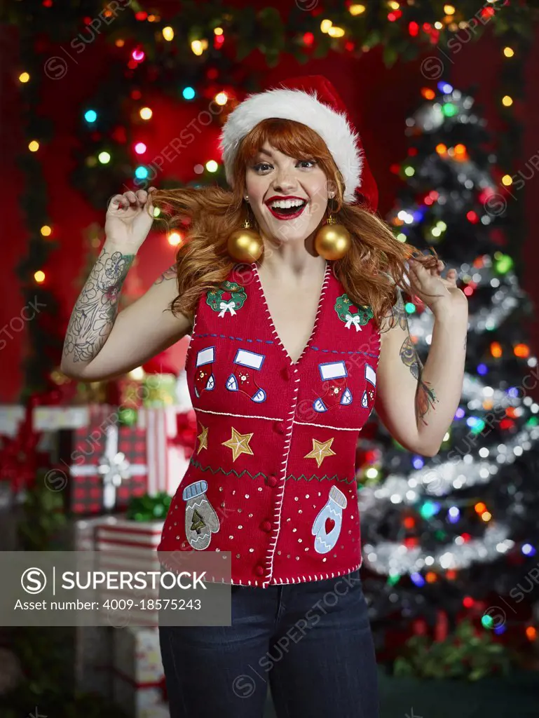 Portrait of red-headed woman with tattoos in an "ugly" Christmas vest making a face and plying with her hair - SuperStock