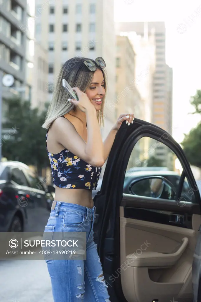 young wealthy Hispanic woman wearing floral top with torn blue jeans, getting into car on busy city street .