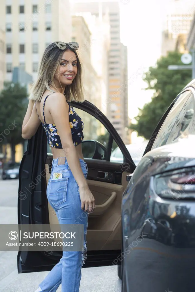 Portrait of Hispanic woman wearing floral top with torn blue jeans, getting into car on busy city street 