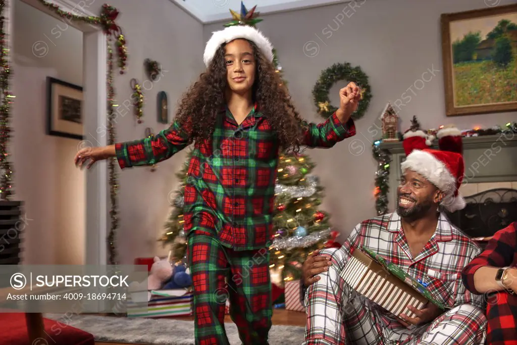 Young girl in Christmas pajamas running towards camera as her father looks on laughing. 