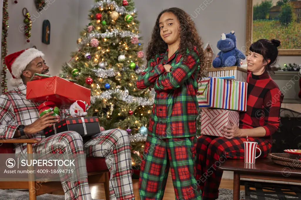 Portrait of a young girl looking into camera with arms crossed, parents in background with piles of gifts on their laps.