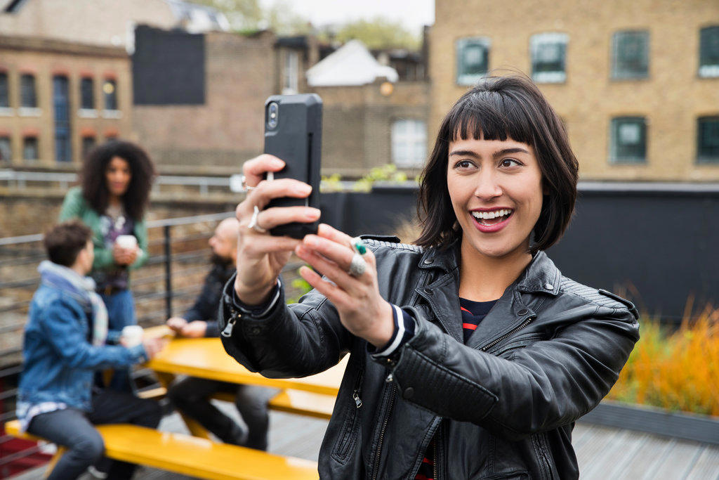 Portrait of Woman taking selfie with group of friends working on tablets at outdoor patio table in co-working space in the background