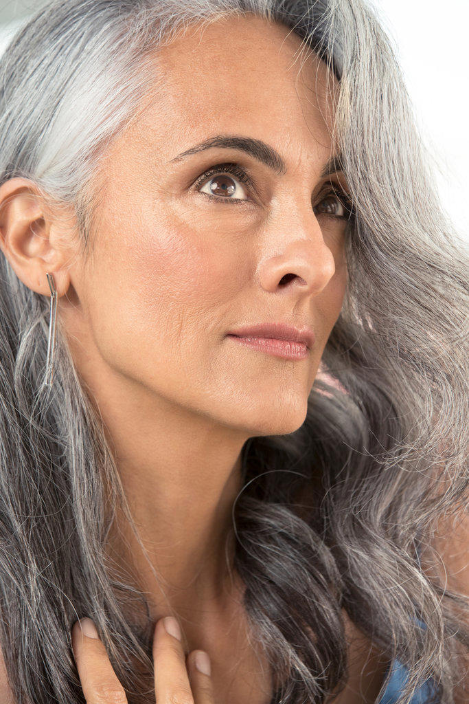 Close up of a middle-aged woman with gray hair, looking up off camera and brushing away her hair.