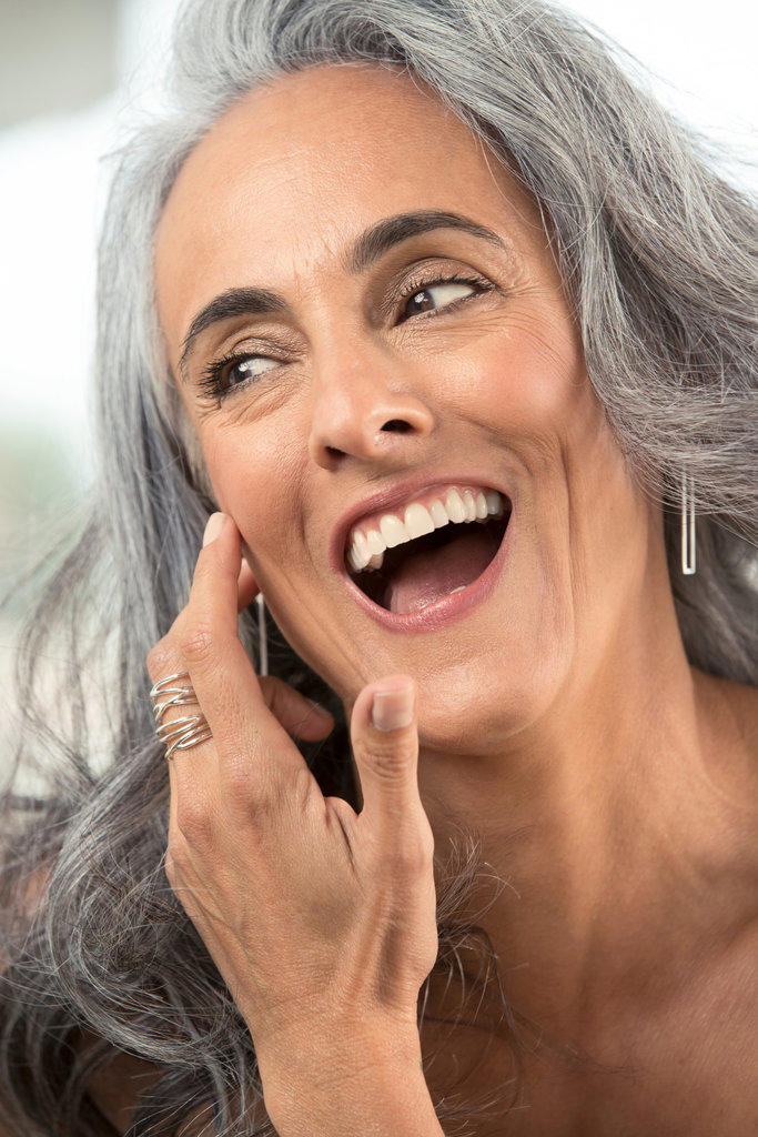 Middle-aged woman with gray hair brushes hair out of her face and looks off camera laughing.