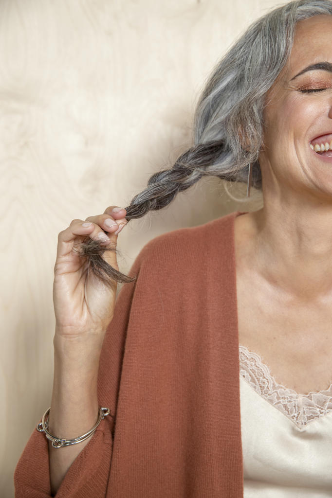 Close up shot of a middle-aged woman with gray hair holding her pony tail off to the side, laughing.