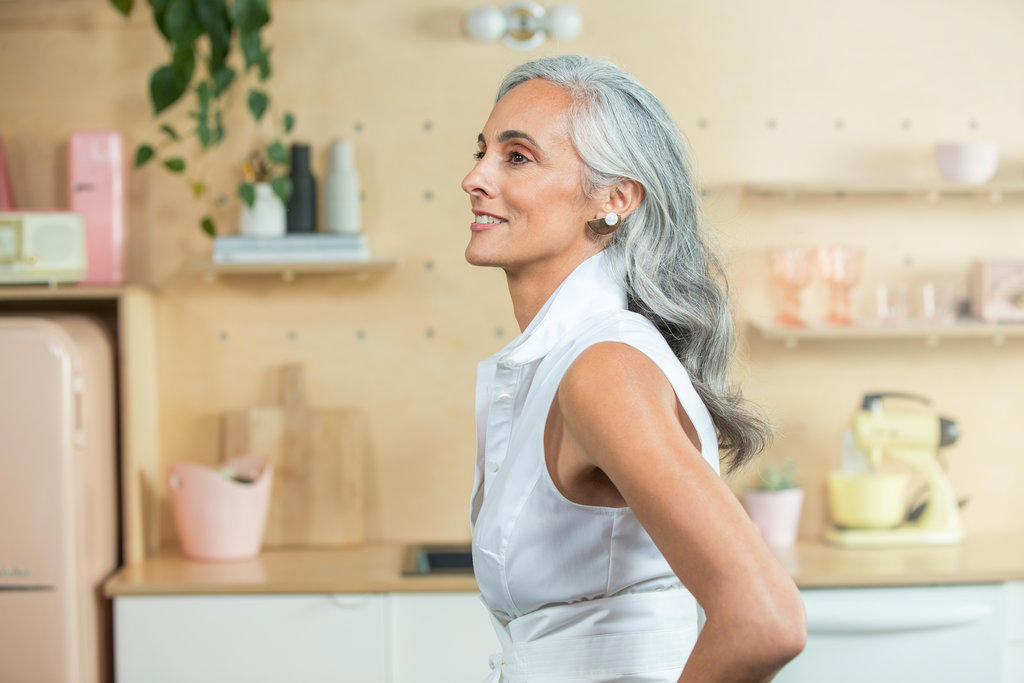 Standing in a  modern kitchen, A youthful middle-aged woman with gray hair looks off camera.