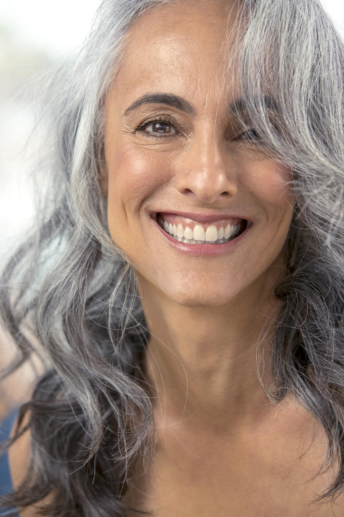 A youthful looking woman with grey hair, looks into camera smiling.