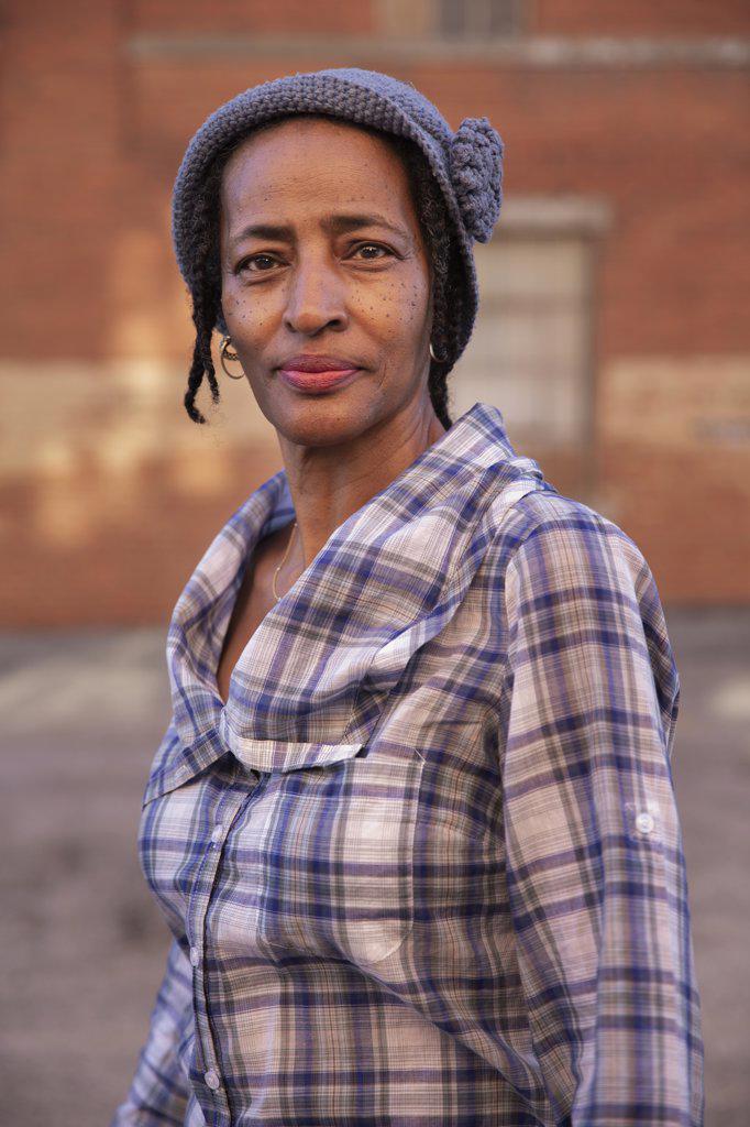 Portrait of older woman wearing knit hat standing in alley, brick wall in background  