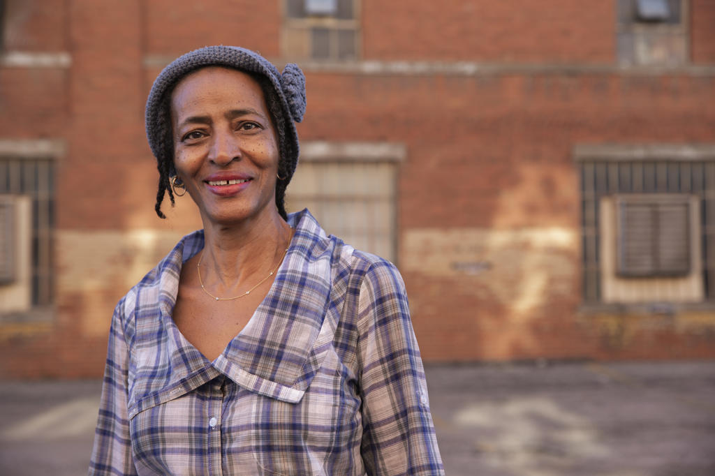 Portrait of older woman wearing knit hat standing in alley smiling looking at camera, brick wall in background  