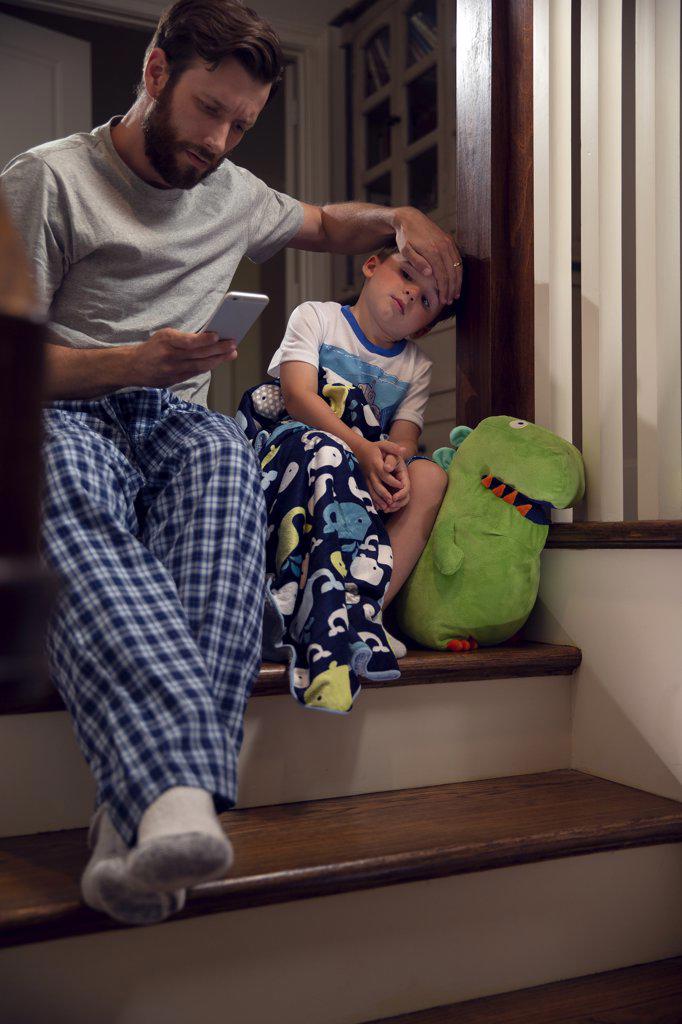 Father and son sitting at top of stairs in home getting ready for bedtime , dad feeling for fever and checking cellphone 