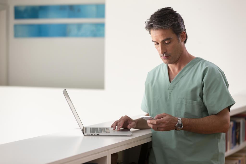 Hispanic Male doctor in his home practicing Tele-medicine, using laptop and cell phone