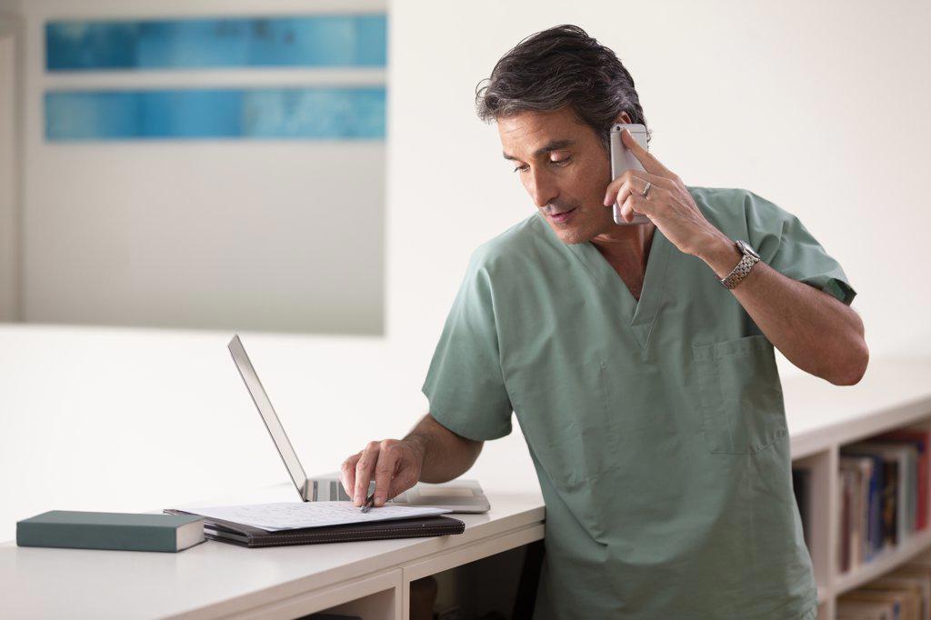 Hispanic Male doctor in his home practicing Telemedicine, talking on cell phone and taking notes 
