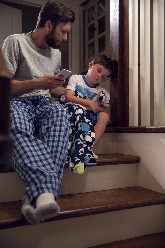 Father and son sitting at top of stairs in home getting ready for bedtime, dad checking on his son who is feeling ill 