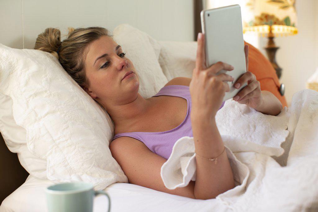 Young woman in bed debating getting up, using tablet to check in with doctor 