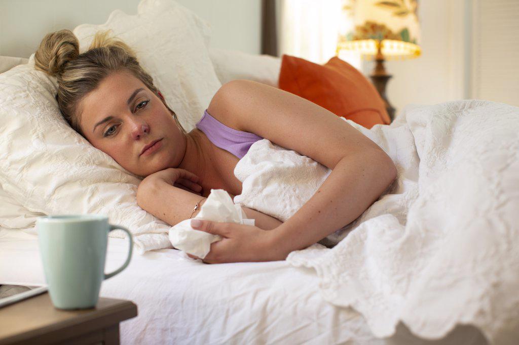 Young woman in her bed debating getting out of bed, holding tissue in hand, smartphone and coffee mug sitting on bedside table 