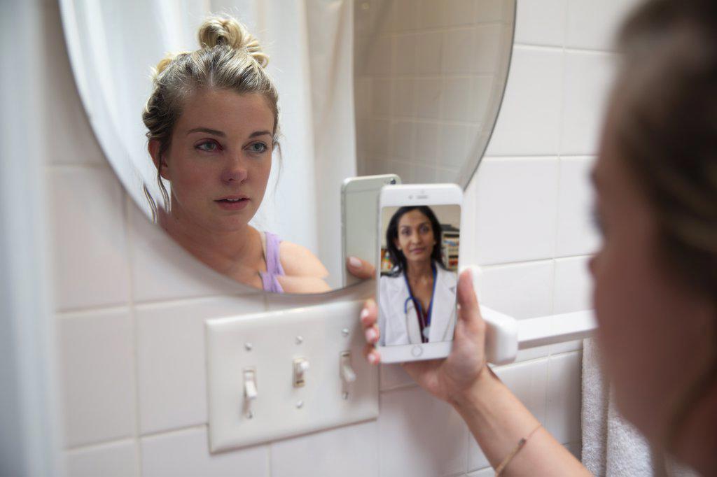 Young woman in her bathroom looking at herself in mirror while talking to doctor on video call