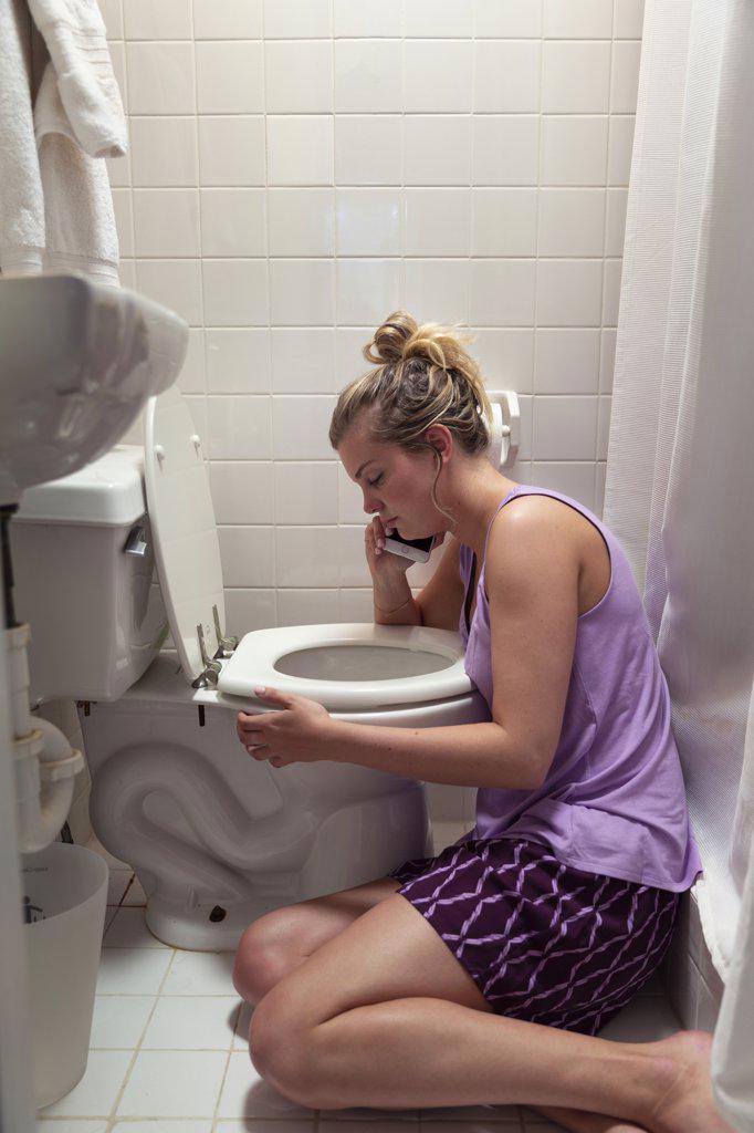 Young woman on the floor in her bathroom feeling sick next to toilet, talking to doctor on cellphone 
