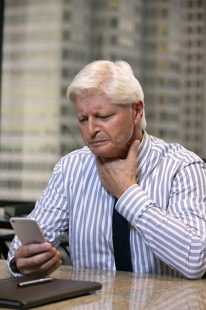 Mature Caucasian man with sore throat fighting off a cold in office of high-rise building downtown, looking at smartphone having a tele-medicine video call with doctor 