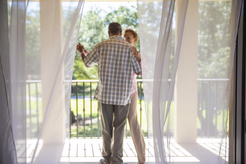 Older couple dancing on porch overlooking  green lawn , seen through sheer curtains , focus on sheers