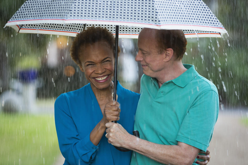 Older couple walking through park with umbrella in the rain, woman laughing and smiling towards camera  