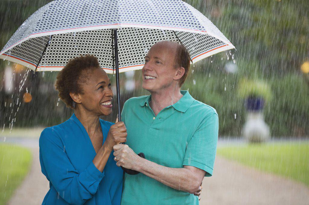 Older couple walking through park with umbrella in the rain, lovingly looking at one another  