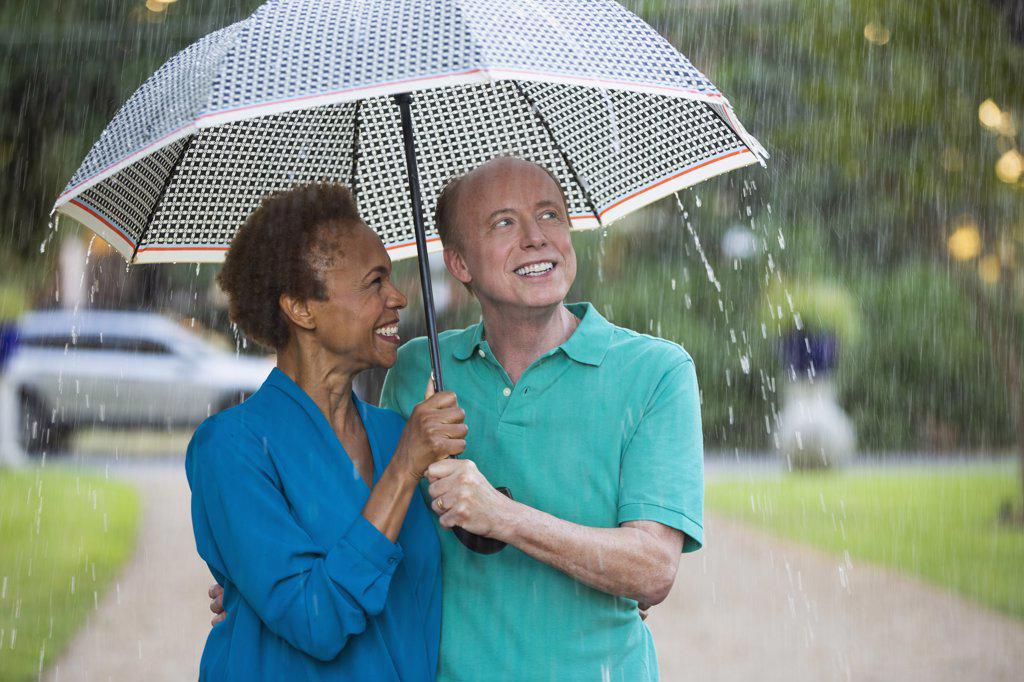 Older couple walking through park with umbrella in the rain, man looking out at the weather 