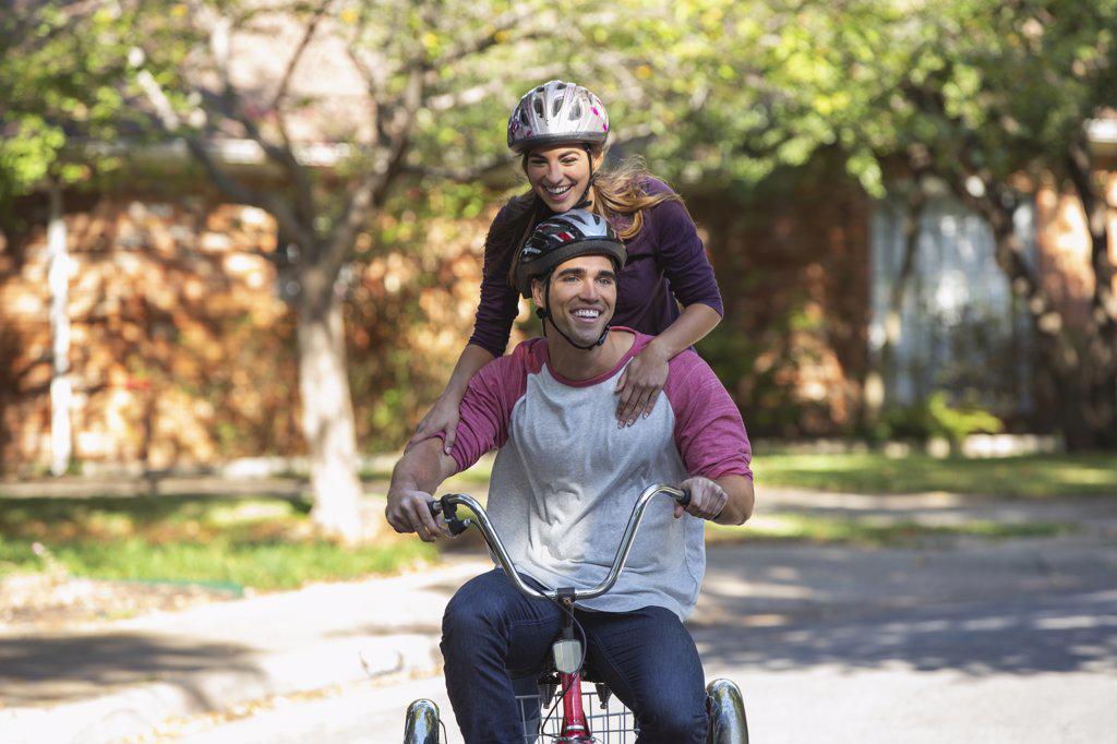 Young couple with helmets riding large tricycle through neighborhood, girl standing on back with her arms on boyfriends shoulders 