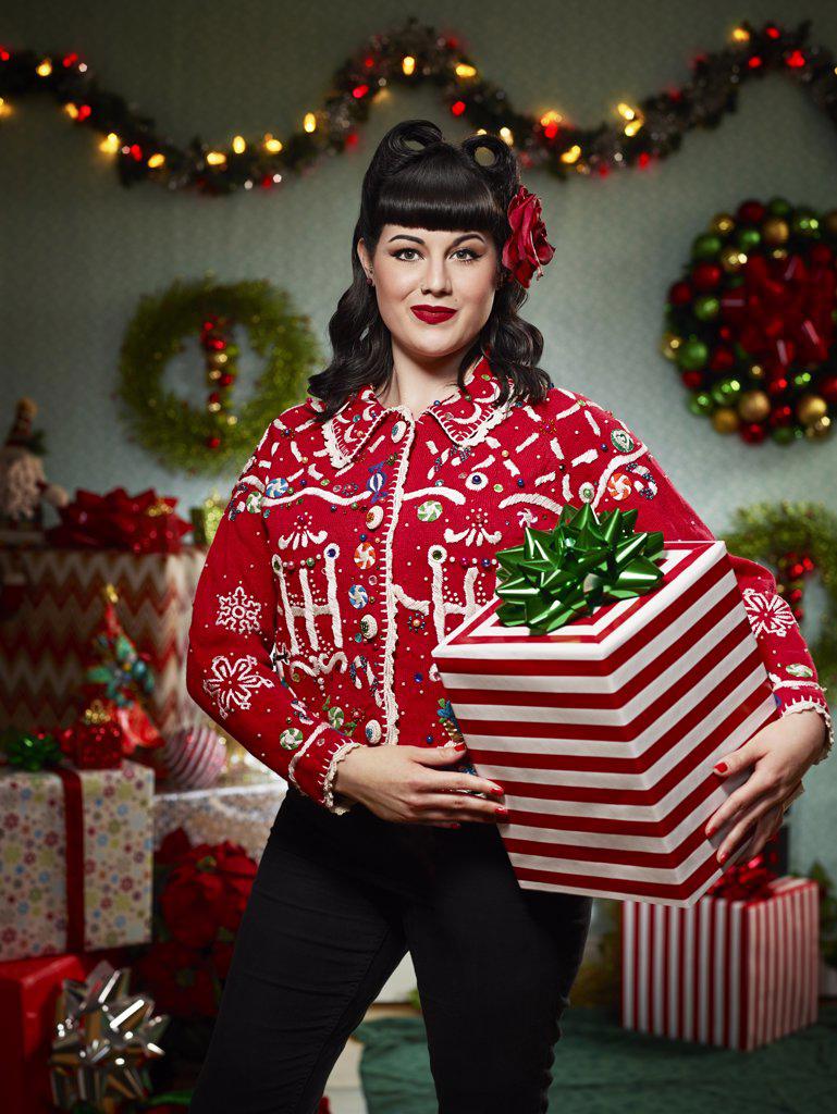 Portrait of woman with a retro hair style, in an "ugly" Christmas sweater holding a present looking at the camera.