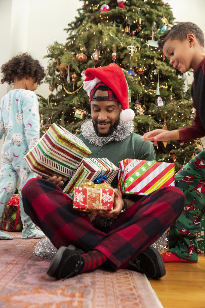A father playing with his kids on Christmas Eve as they pile presents on his lap.