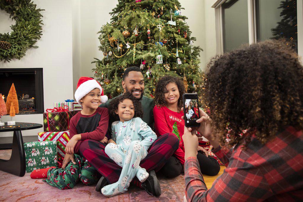 Young Black family taking pictures on mobile phone under Christmas tree.
