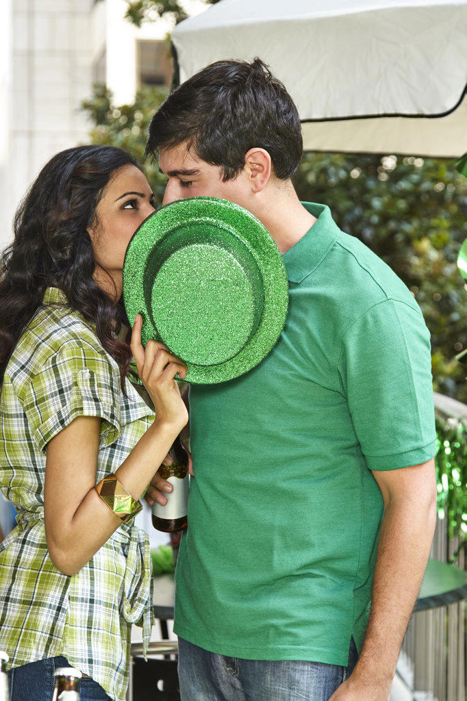 Young diverse couple kissing behind a green St. Patrick's Day hat while at party on balcony. 