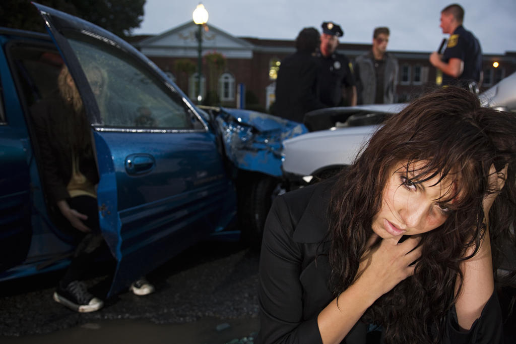 Injured young woman sitting in street  with scene of an automobile accident behind her 