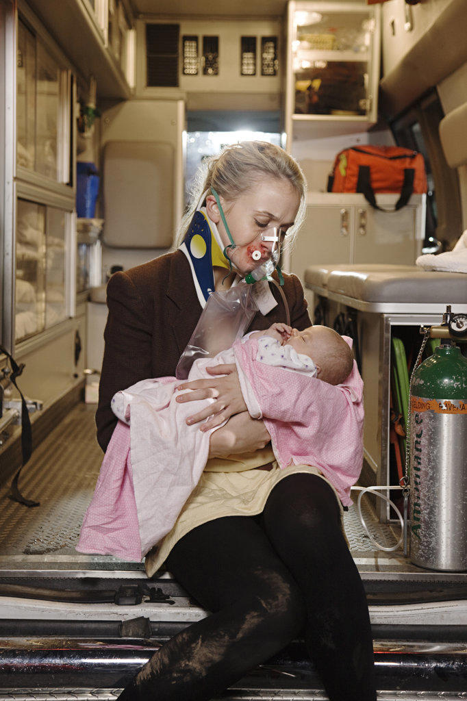 Portrait of injured young woman wearing oxygen mask holding infant sitting  on back of an open ambulance 