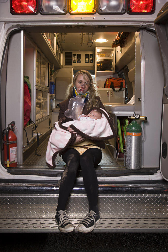 Portrait of injured young woman wearing oxygen mask holding and infant looking at camera  sitting  on back of an open ambulance 